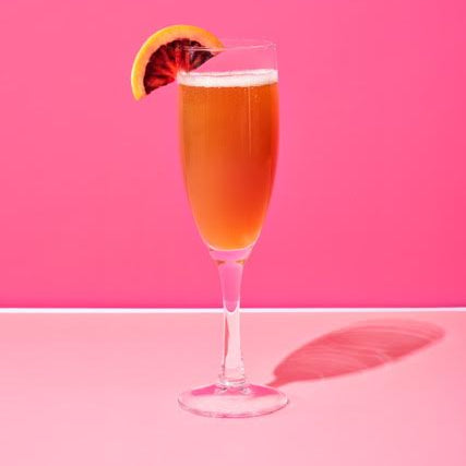 Champagne flute with an amber cocktail garnished with a blood orange slice against a pink background.
