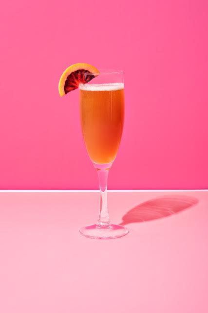 Champagne flute with an amber cocktail garnished with a blood orange slice against a pink background.