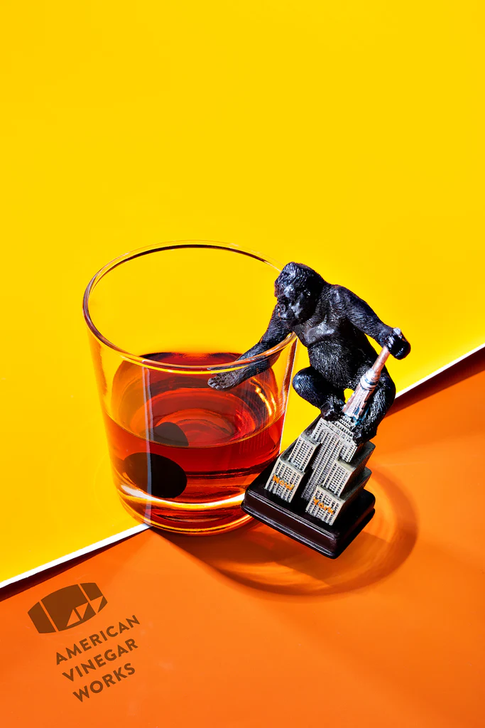 Toy gorilla on a miniature Empire State Building beside a glass of amber vinegar; AMERICAN VINEGAR WORKS logo visible
