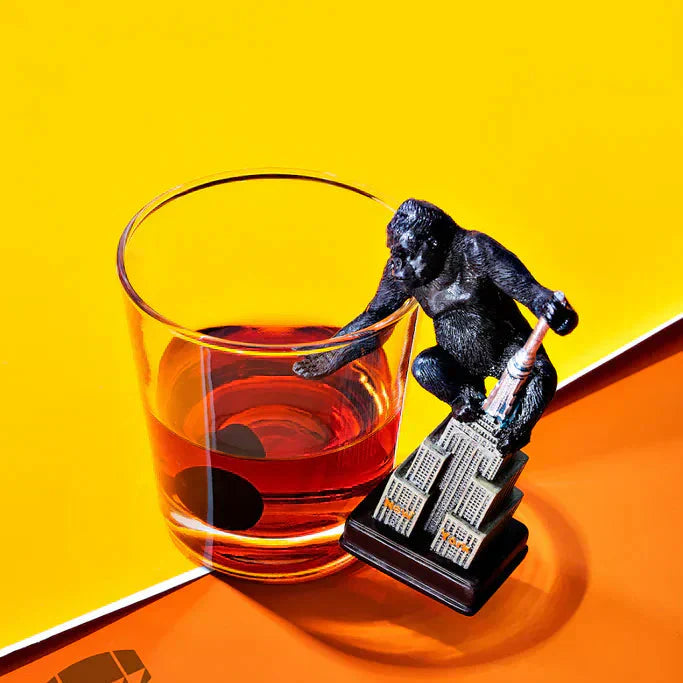 Toy gorilla perched on a mini Empire State Building next to an amber cocktail on yellow background