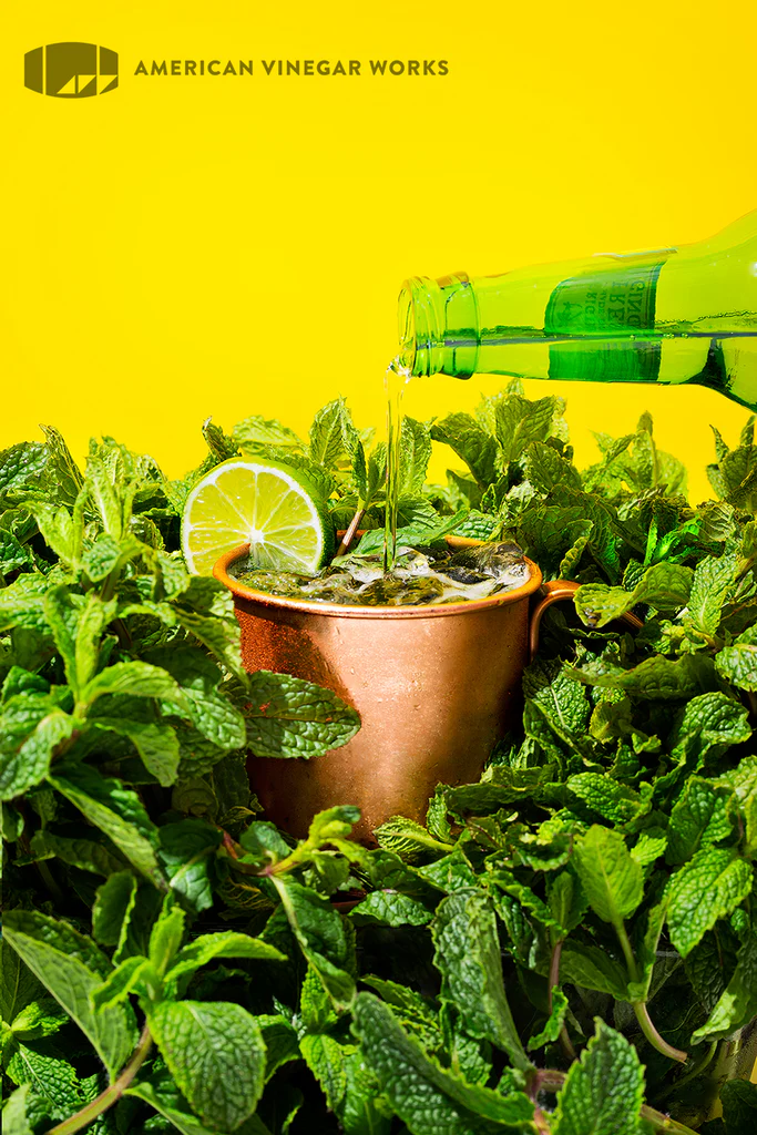 Copper mule mug with mint, lime slice and green bottle pouring over ice; American Vinegar Works logo at top.