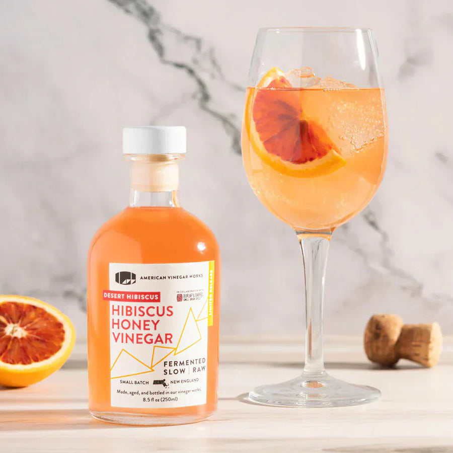 Hibiscus Honey Vinegar bottle with white cap next to rosé spritz garnished with blood orange