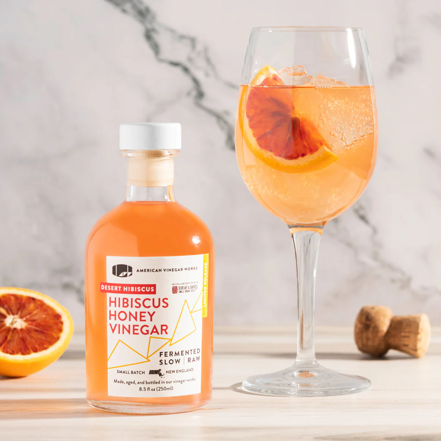 Bottle of Hibiscus Honey Vinegar with label visible beside an orange hibiscus spritz in a wine glass on marble