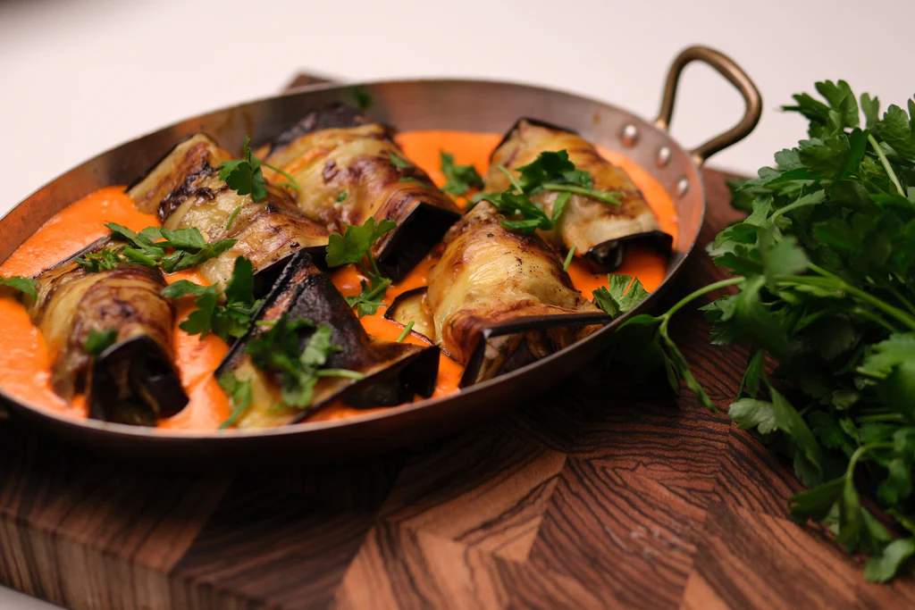 Rolled eggplant stuffed with goat cheese resting in roasted red pepper sauce in a copper skillet on a wooden board, garnished with parsley