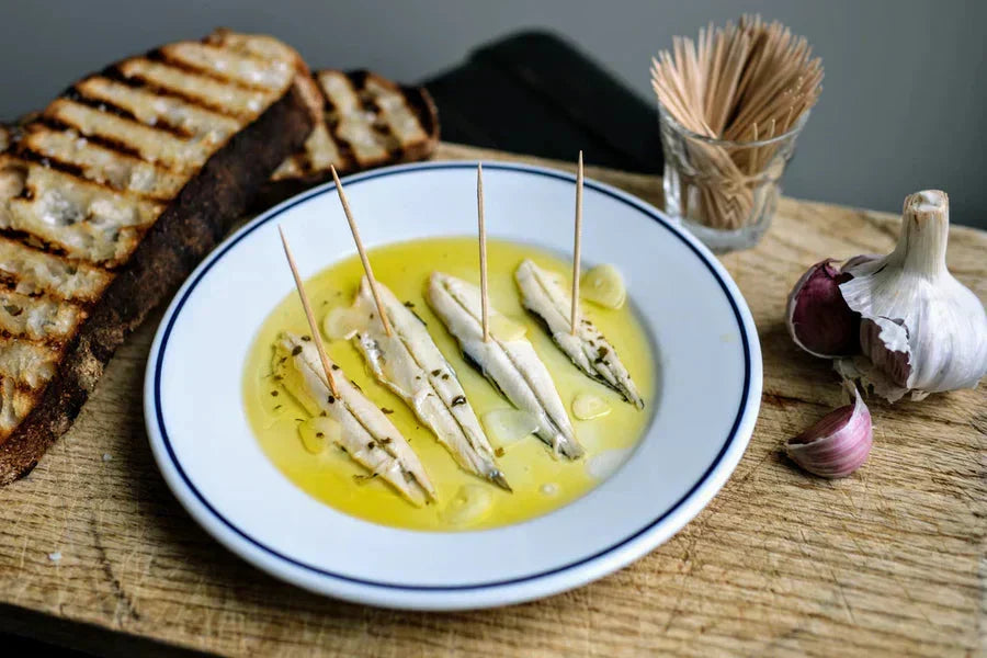 Marinated white anchovies in oil with toothpicks, grilled bread, and garlic on a wooden board bowl