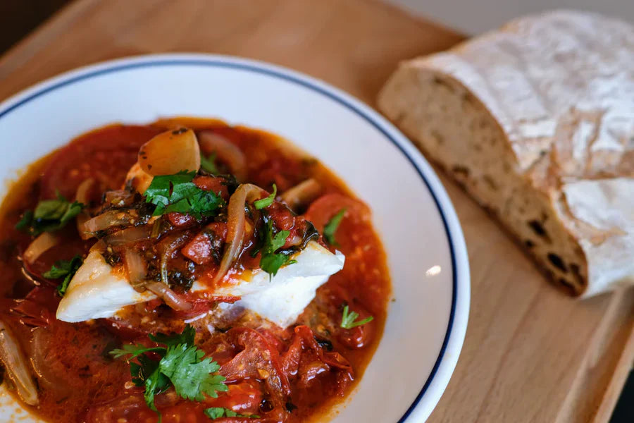 Roasted white fish topped with stewed tomatoes, onions, and cilantro in a white bowl with blue rim, bread on wooden board