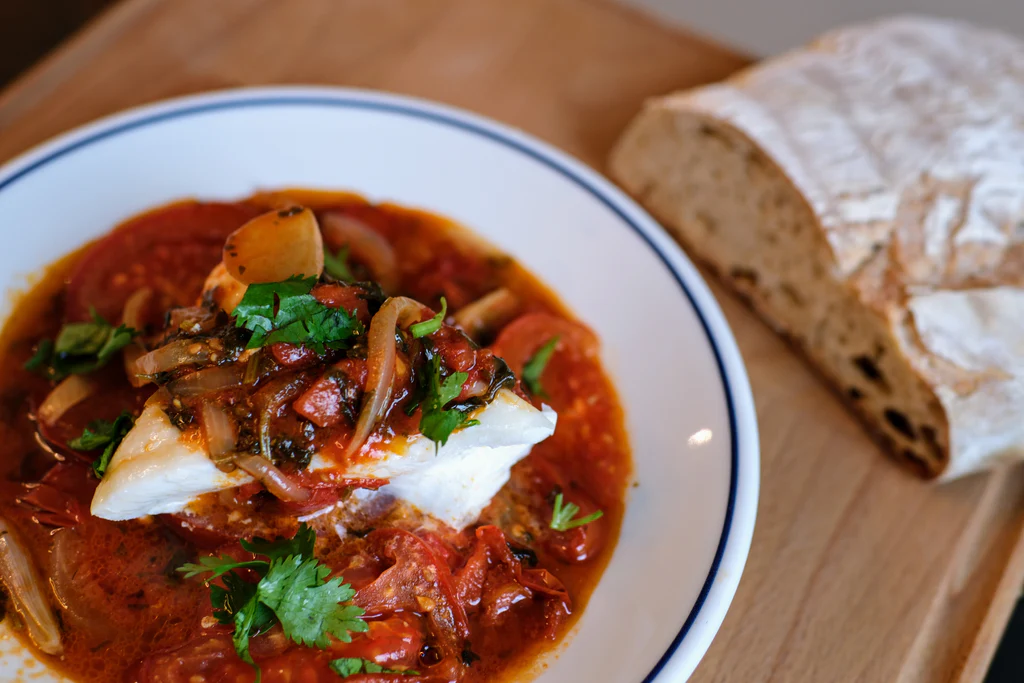Roasted white fish on stewed tomatoes and onions, with cilantro and crusty bread on a wooden board
