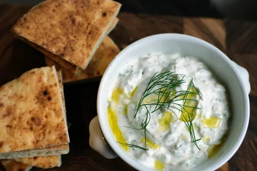 Overhead view of tzatziki in white bowl with olive oil drizzle and pita on wooden board