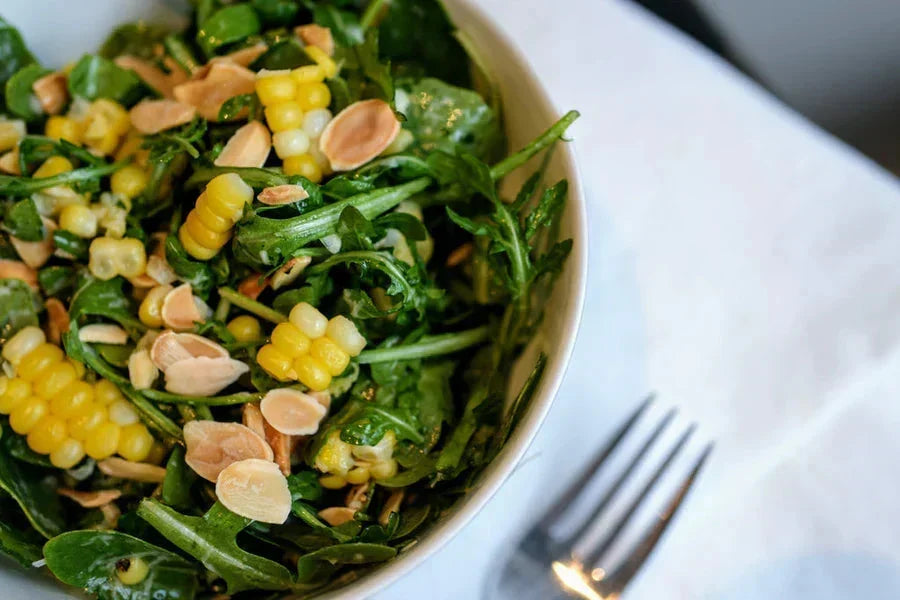 Bowl of arugula and corn salad with lemon-oil dressing, almonds, and fork on a white napkin