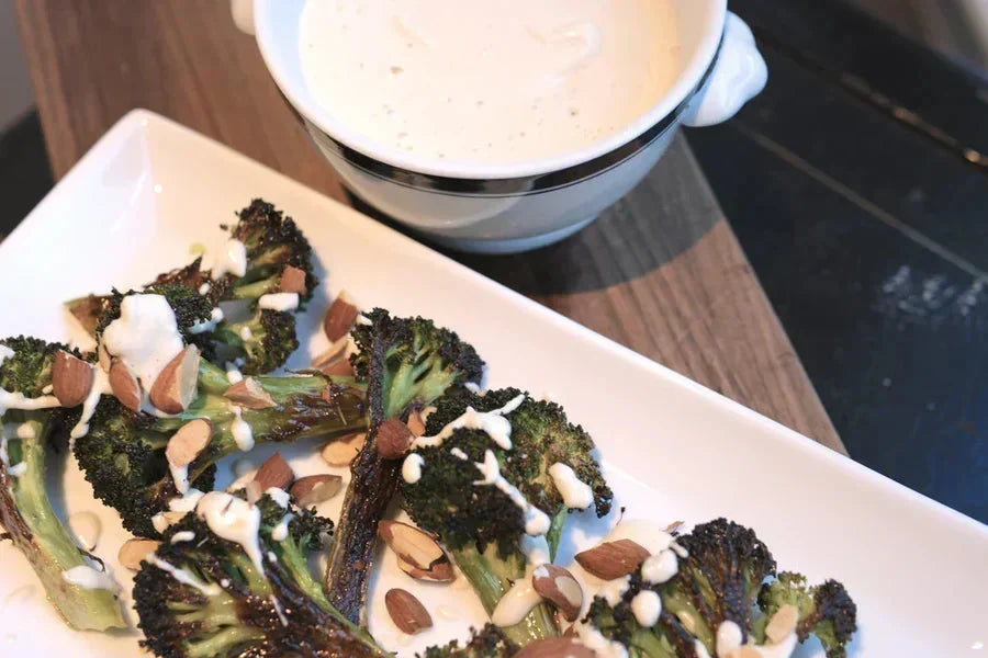 Roasted broccoli with cashew cream sauce and chopped almonds on a white platter beside a sauce bowl