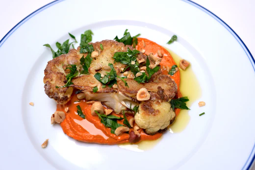 Roasted cauliflower steak on romesco sauce, garnished with chopped hazelnuts and parsley on a white rimmed plate.