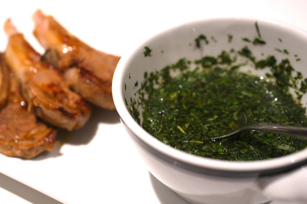 Bowl of chopped fresh mint sauce with a spoon beside pan-fried lamb chops on a white surface