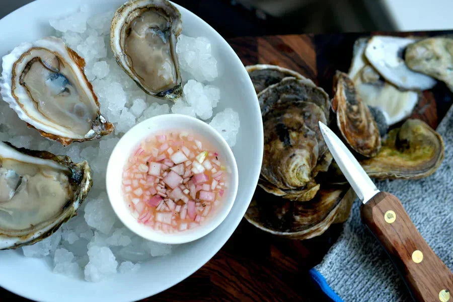 Pink mignonette bowl with shallots and shucked oysters on crushed ice with a knife on wooden board