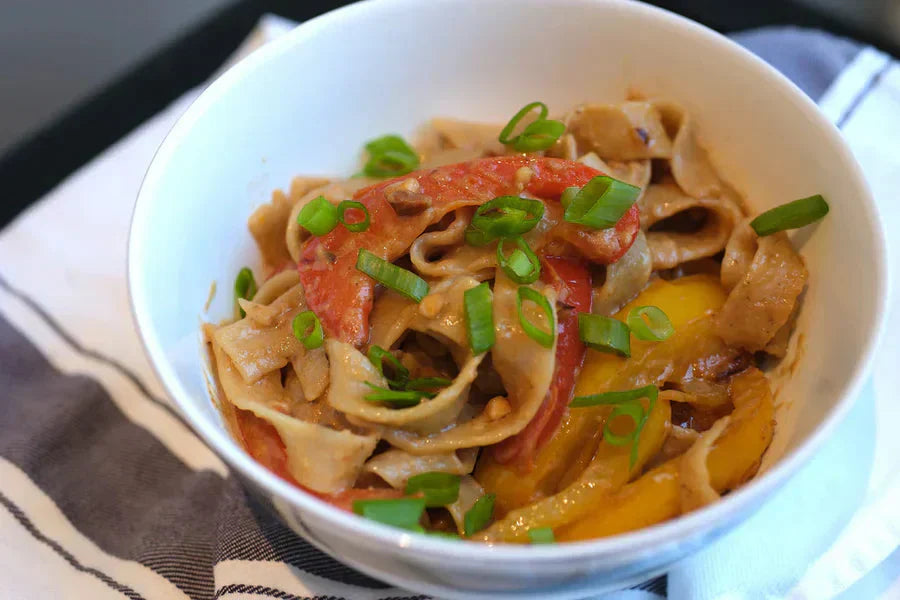 Bowl of peanut sauce noodles with sliced red, yellow bell peppers and scallions on a striped napkin.