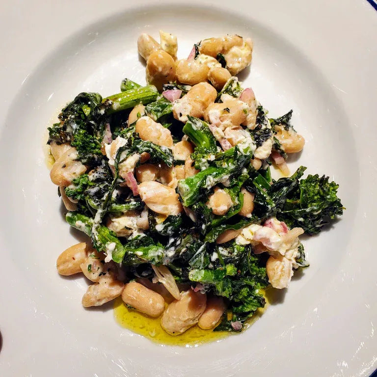 A white rimmed plate filled with grilled broccoli rabe salad, white beans, and goat cheese.