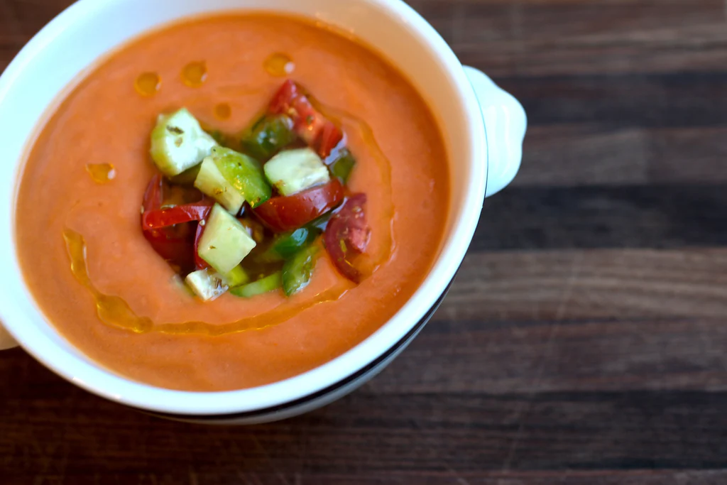 Bowl of chilled gazpacho with diced cucumber, tomatoes, green pepper, and olive oil on wooden board