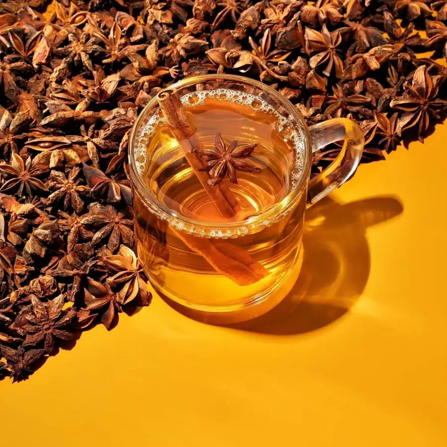 Clear mug of amber spiced liquid with cinnamon stick and star anise, pile of star anise on yellow background