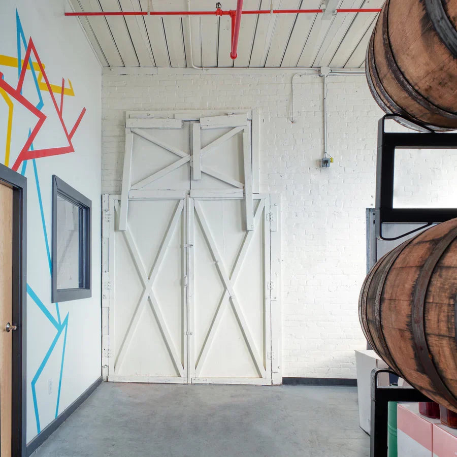Vinegar workshop interior: wooden barrels stacked on racks beside white barn doors and a colorful geometric wall mural.