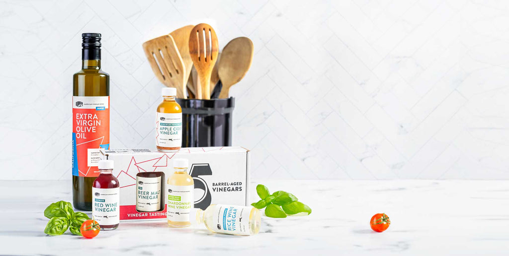 Front of vinegar tasting kit and olive oil with five vinegar bottles, basil and cherry tomatoes on marble background