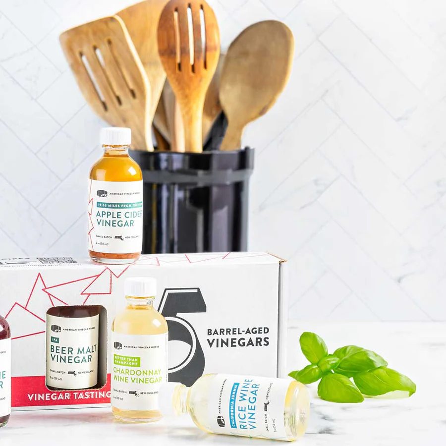 Vinegar tasting gift set with olive oil & apple cider bottles, staged with basil & wood utensils.