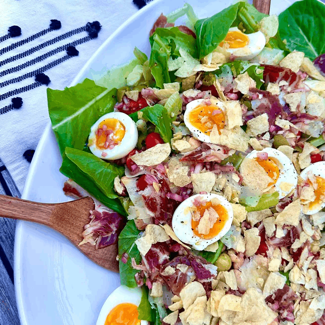 Top-down view of Spanish tapas salad with jamón, quail eggs, Manchego, chips, & wooden spoons at rim