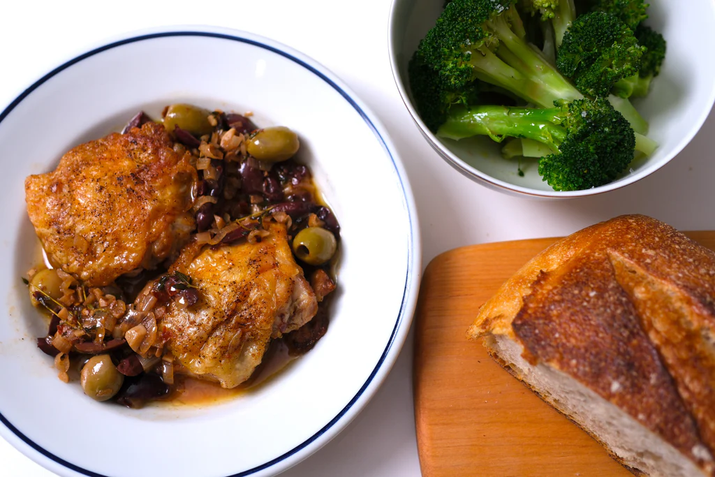 Pan-seared chicken thighs braised with olives and onions in white bowl, with broccoli, crusty bread