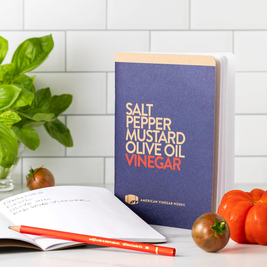 A blue notebook titled 'Salt Pepper MUSTARD OIL Vinegar' with a pen and some tomatoes on its surface, placed on a kitchen counter with a plant in the background.