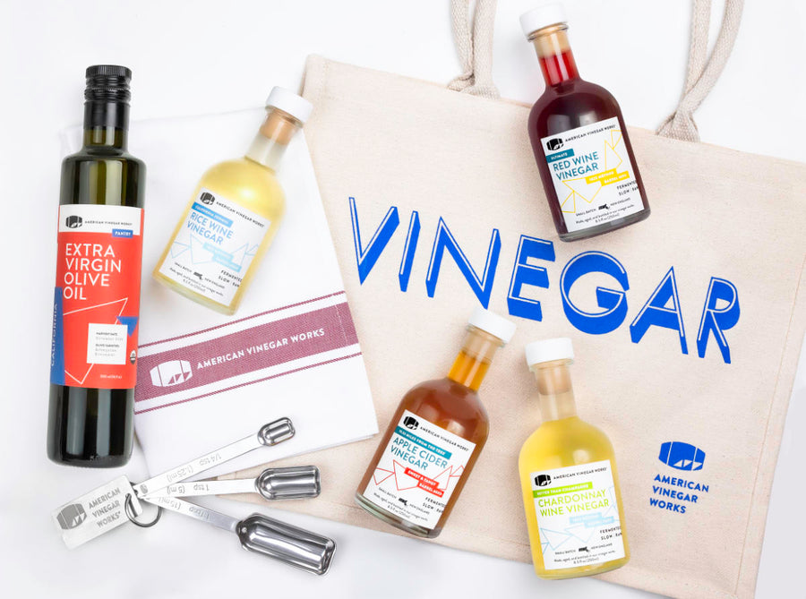 A  bundle from American Vinegar Works featuring a branded "VINEGAR" tote bag, a bottle of Extra Virgin Olive Oil, and four small bottles of specialty vinegars including Rice Wine, Red Wine, Apple Cider, and Chardonnay Wine vinegar, accompanied by a branded tea towel and a set of measuring spoons