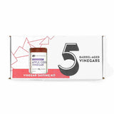 A white rectangular product box for an American Vinegar Works "5 Barrel-Aged Vinegars" tasting kit, featuring a cutout window displaying a bottle of Maine Blueberry Apple Cider Vinegar and stylized red geometric line art.
