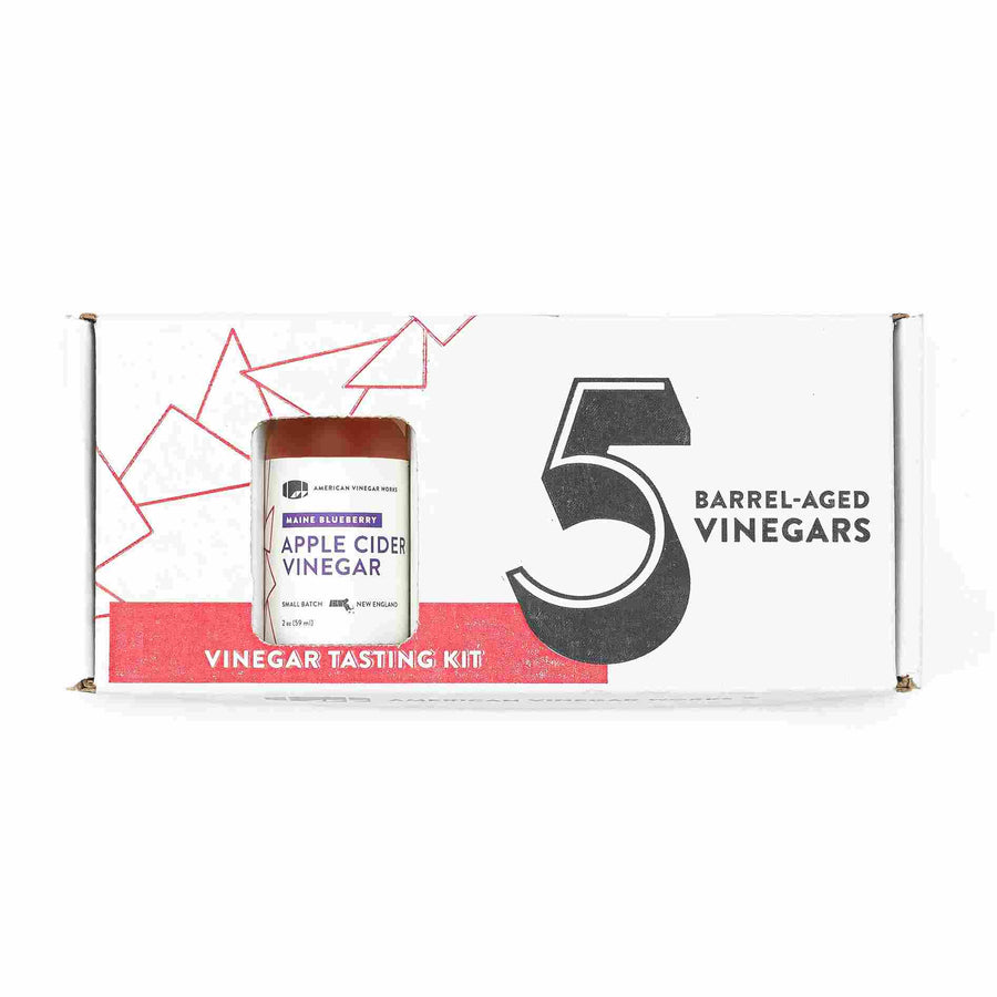 A white rectangular product box for an American Vinegar Works "5 Barrel-Aged Vinegars" tasting kit, featuring a cutout window displaying a bottle of Maine Blueberry Apple Cider Vinegar and stylized red geometric line art.