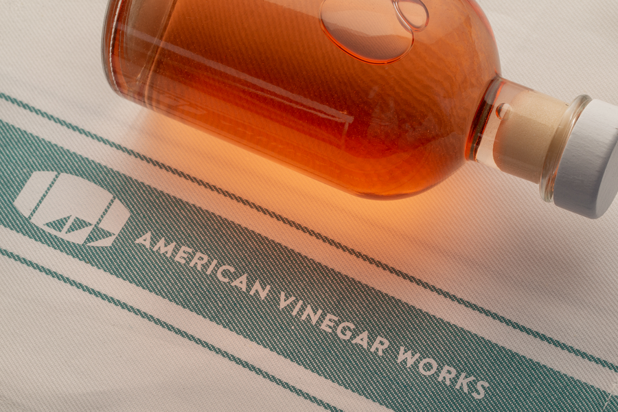 Amber vinegar bottle resting on a white 100% cotton tea towel with teal stripes and ‘American Vinegar Works’ logo