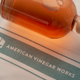 Amber vinegar bottle resting on a white 100% cotton tea towel with teal stripes and ‘American Vinegar Works’ logo