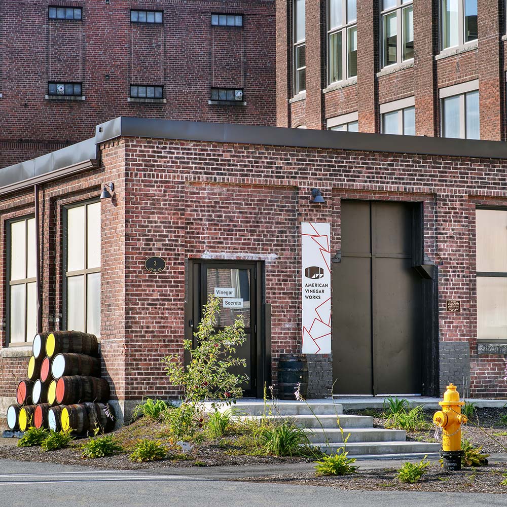Brick building entrance with sign 'AMERICAN VINEGAR WORKS', door labeled 'Vinegar Secrets', stacked barrels, yellow fire hydrant, plaque '3'.