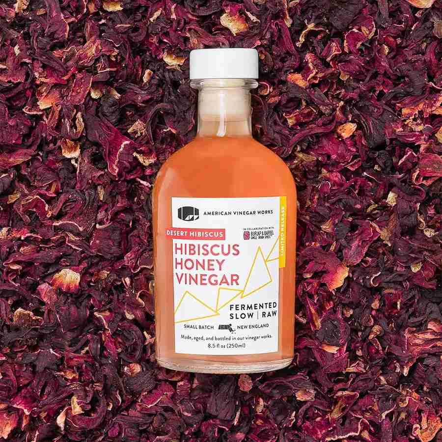 A bottle of American Vinegar Works Desert Hibiscus Honey Vinegar centered on a bed of dried hibiscus flowers.