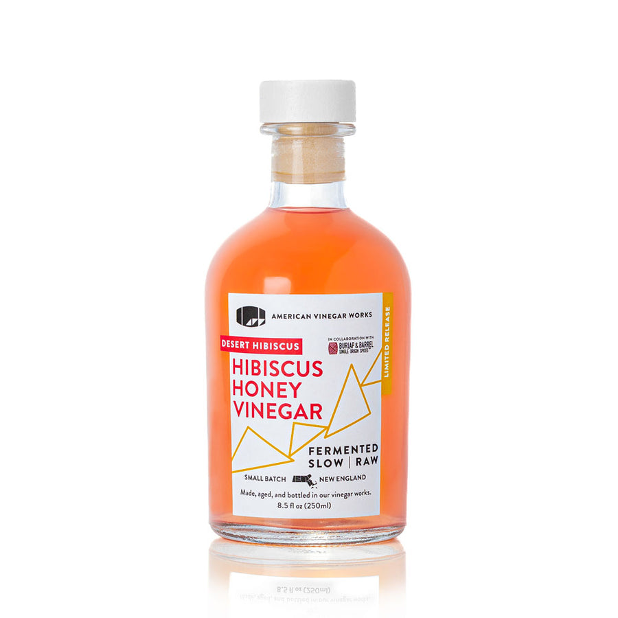 A bottle of American Vinegar Works Desert Hibiscus Honey Vinegar, a limited release in collaboration with Burlap & Barrel, featuring a white cap and vibrant pinkish-orange liquid.