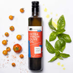 A bottle of American Vinegar Works Extra Virgin Olive Oil surrounded by fresh cherry tomatoes, basil leaves, salt crystals, and droplets of oil on a white background.