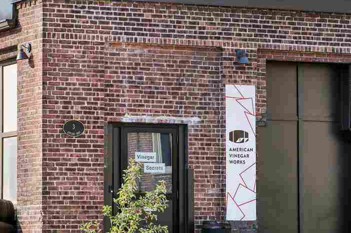 Exterior view of brick American Vinegar Works facility with an entrance sign & stacked wooden barrel