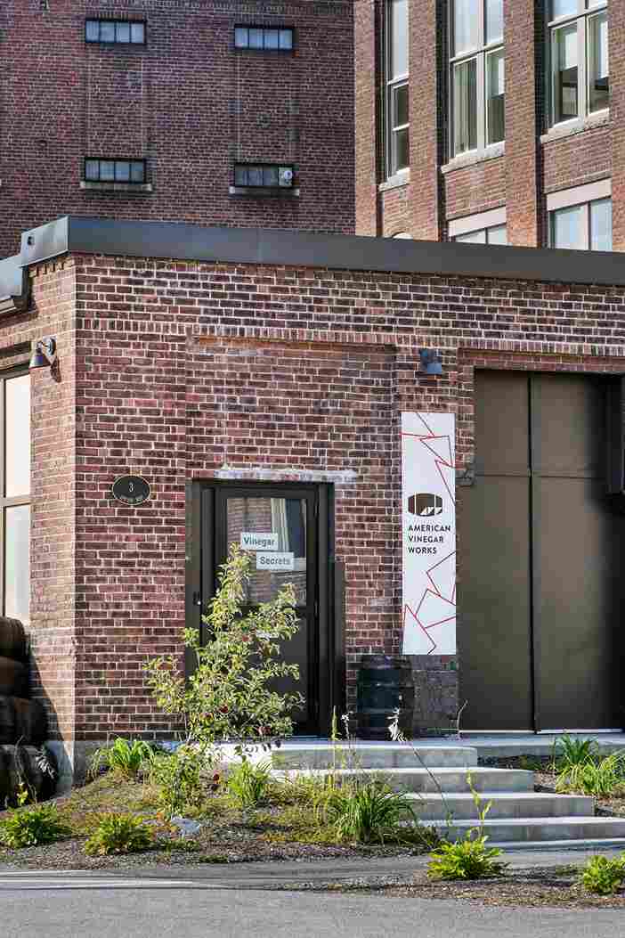Exterior view of brick American Vinegar Works facility with an entrance sign & stacked wooden barrel