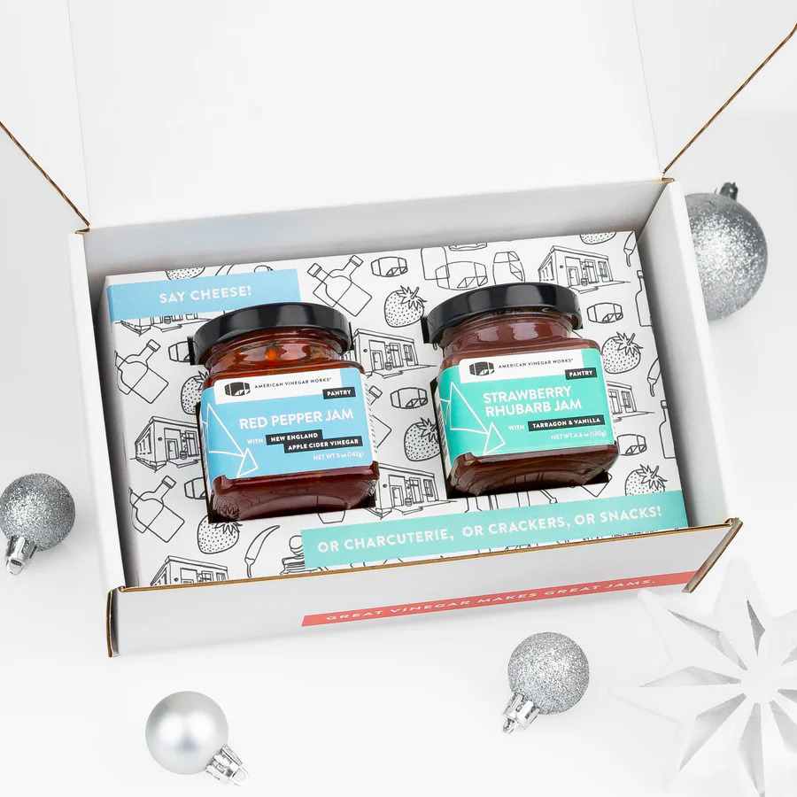 Open white gift box with Red Pepper & Strawberry Rhubarb jam jars, silver ornaments, and snowflakes.