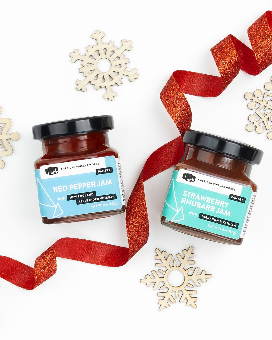 Two glass jars of Red Pepper Jam and Strawberry Rhubarb Jam with red ribbons and wooden snowflakes.