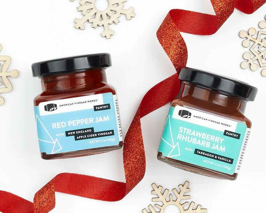 Two glass jars of Red Pepper Jam and Strawberry Rhubarb Jam with red ribbons and wooden snowflakes.