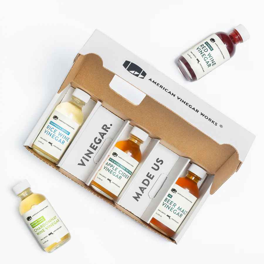 An open American Vinegar Works gift box containing three small bottles of vinegar, with two additional bottles of Red Wine Vinegar and Chardonnay Wine Vinegar placed outside the box on a white background.