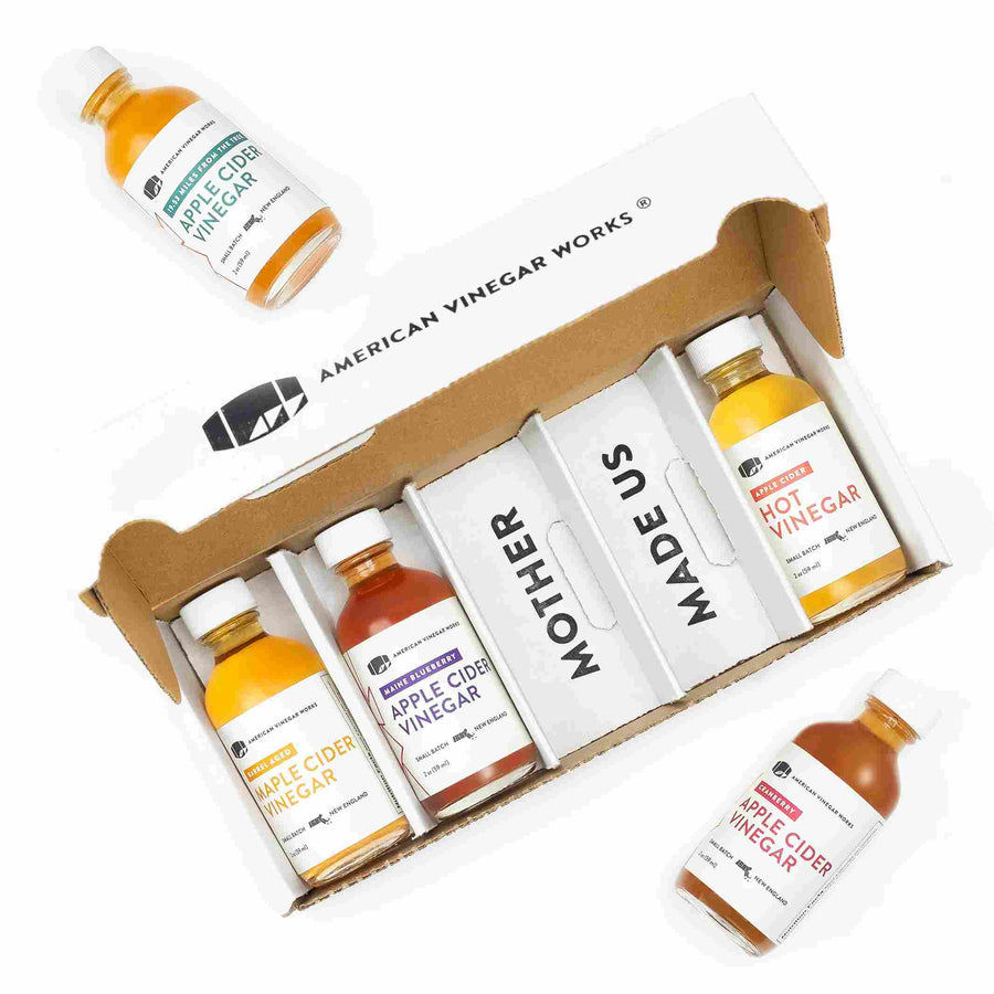 A gift box set from American Vinegar Works containing four small glass bottles of specialty vinegars labeled Maple Cider Vinegar, Maine Blueberry Apple Cider Vinegar, Hot Vinegar, and Apple Cider Vinegar, with an additional bottle of Apple Cider Vinegar placed outside the box. The box insert reads "MOTHER MADE US".