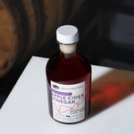 Angled shot of Blueberry Apple Cider Vinegar bottle on a white surface with a dark barrel background