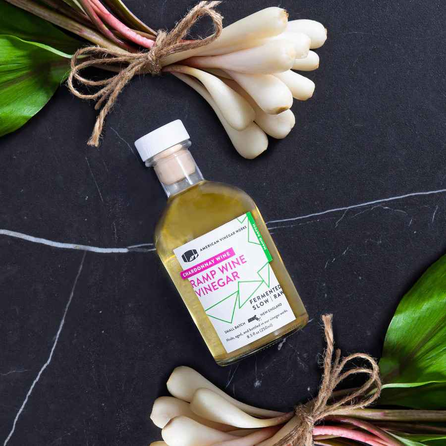 A top-down flat lay view of an 8.5 fluid ounce bottle of American Vinegar Works Chardonnay Wine Ramp Wine Vinegar resting on a black marbled background, flanked by two bundles of fresh wild ramps tied with twine.