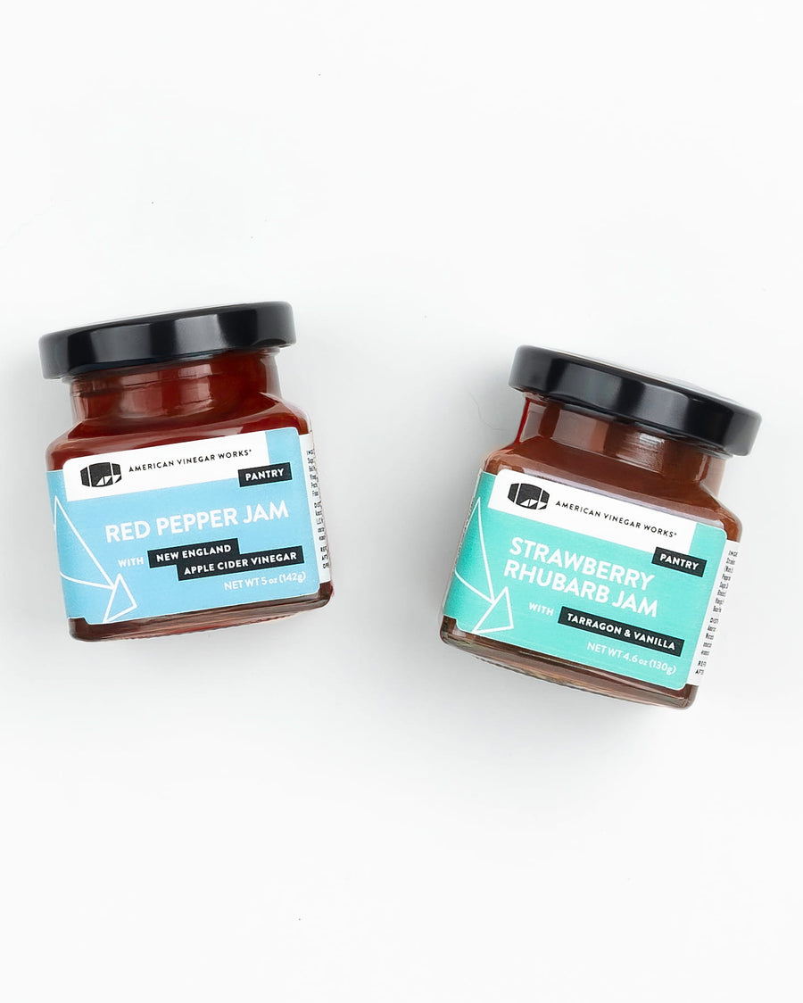 A square-cropped top-down view of two jars from the American Vinegar Works Jam Gift Set, featuring Red Pepper Jam and Strawberry Rhubarb Jam side by side against a white background.