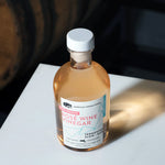 An angled, high-angle shot of an 8.5 fluid ounce bottle of American Vinegar Works Pink Pacific Rosé Wine Vinegar sitting on a white surface, casting a warm shadow with a wooden barrel visible in the dark background.