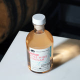 An angled, high-angle shot of an 8.5 fluid ounce bottle of American Vinegar Works Pink Pacific Rosé Wine Vinegar sitting on a white surface, casting a warm shadow with a wooden barrel visible in the dark background.