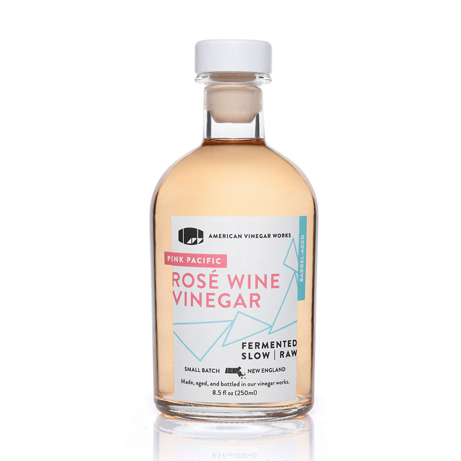 The front view of an 8.5 fluid ounce bottle of American Vinegar Works Pink Pacific Rosé Wine Vinegar, showcasing a light pink liquid and a white label with pink and blue geometric accents against a clean white background.