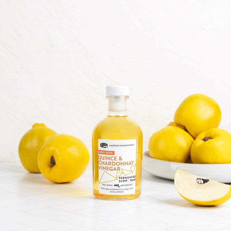 A bright, front lifestyle view of an 8.5 fluid ounce bottle of American Vinegar Works Quince and Chardonnay Vinegar standing on a white tiled surface, surrounded by whole fresh yellow quince fruit and a single cut slice in the foreground.